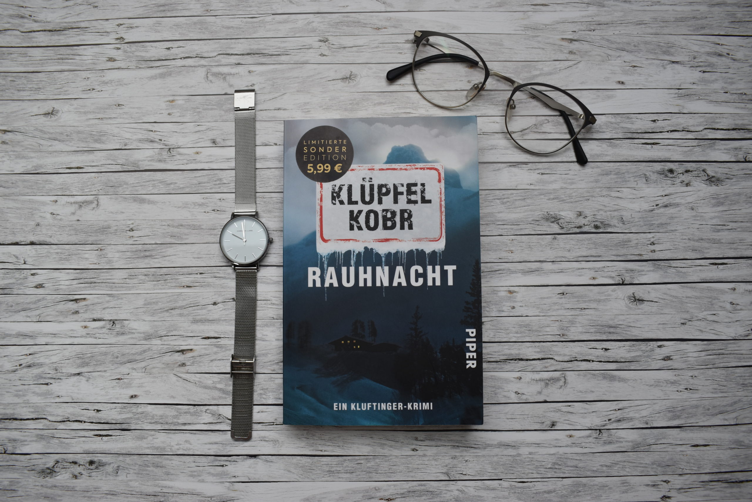 Eine silberne Uhr liegt neben dem Krimi Rauhnacht. Darüber liegt rechts in der Ecke eine Brille.